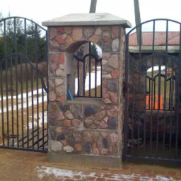 Otwarta brama wjazdowa z czarnego metalu, osadzona w kamiennych słupkach z okienkiem, na ceglanym podjeździe z resztkami śniegu.